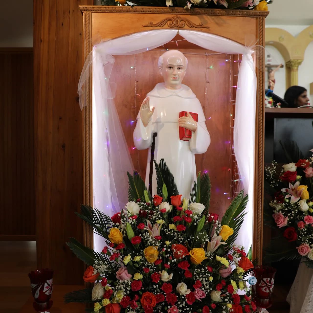 Statue of St. Chavara in a wooden shrine draped with white tulle and banked with flowers