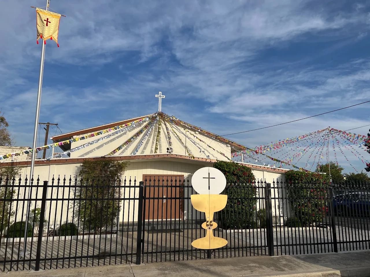 Church with pyramidal roof and cross, draped in festival bunting, with a chalice-and-host cutout at the gate