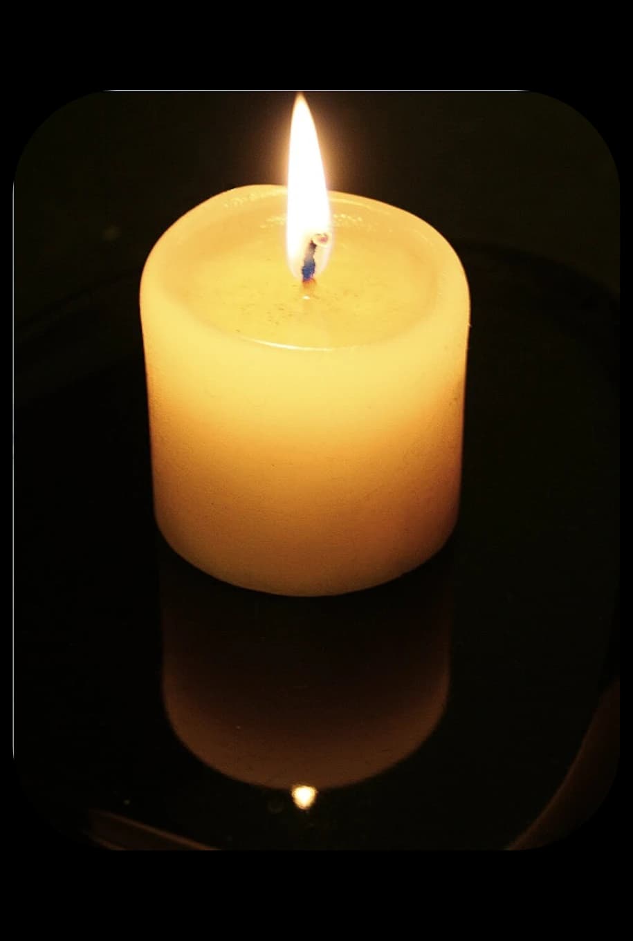 Single lit white pillar candle glowing on a dark reflective surface